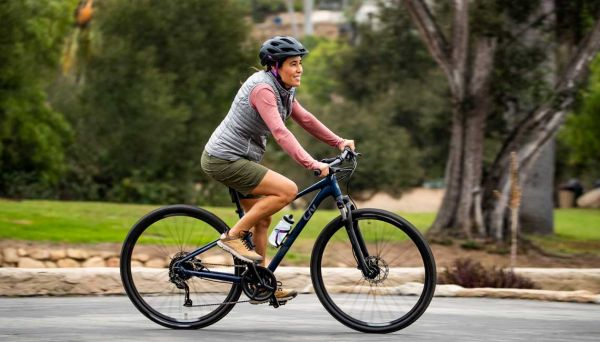  woman of colour cycles a hybrid bike, smiling, along the road. She's wearing shorts, a gilet and a helmet.